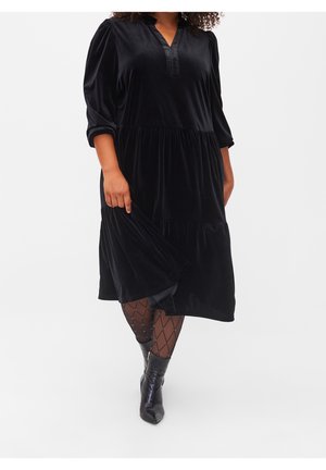 Mujer con un vestido midi de terciopelo negro con volantes recogidos, mangas de tres cuartos, medias estampadas y botas negras hasta el tobillo, caminando hacia adelante.