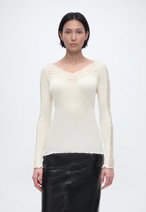 Žena s krátkymi tmavými vlasmi má na sebe priliehavý biely dlhý rukáv s čipkovými detailmi a čiernu koženú sukňu, stojaca na jednoduchom pozadí.