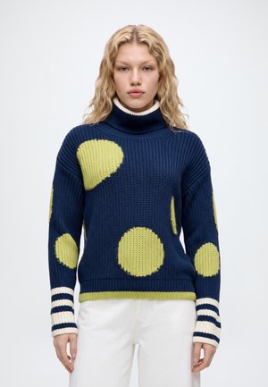 Un mannequin se tient devant un fond uni, vêtu d’un pull bleu marine à cercles vert citron, d’un pantalon et de chaussures blancs, et portant un sac blanc à bandoulière.