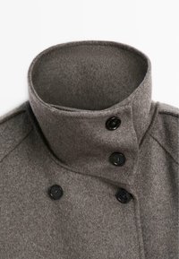 Cappotto di lana grigia con colletto alto, caratterizzato da bottoni neri e una texture morbida, che enfatizza un design strutturato e uno stile minimalista.