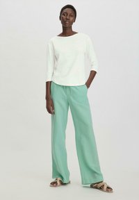 Ein weißes, locker sitzendes 3/4-Ärmel-Oberteil, kombiniert mit mintgrünen, weit geschnittenen Hosen und braunen Sandalen mit Knotendetail.
