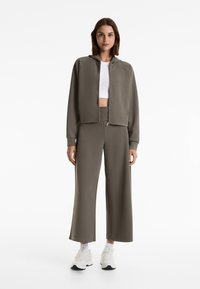 Khakifarbene Kapuzenjacke mit Reißverschluss, geripptem Muster und passenden weit geschnittenen Hosen. Darunter ein weißes Crop-Top und helle Sneakers.