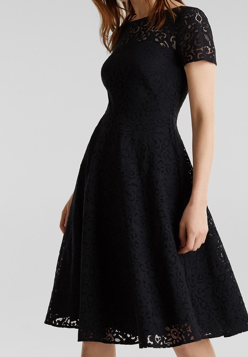 Femme portant une robe en dentelle noire ajustée avec des manches courtes et une jupe évasée, debout devant un fond uni gris clair.
