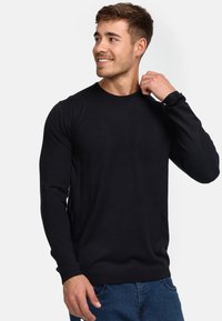 Pull noir à manches longues en tissu lisse, avec un col rond et des poignets côtelés, associé à un jean en denim bleu.
