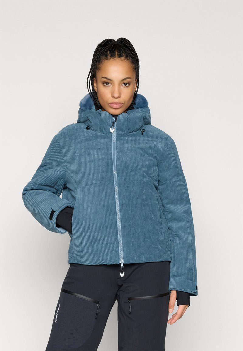 Modrá kordurová bunda s kapucí a zipovým zapínáním, s texturovaným povrchovým zpracováním a elastickými manžetami, nošená přes černé kalhoty.