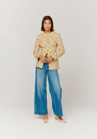 Giacca a quadri gialla con bottoni, dal taglio aderente, abbinata a pantaloni in denim a gamba larga. Il modello indossa tacchi leggeri. Tessuto morbido e texture liscia.