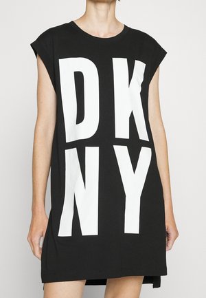 Person iført en sort ærmeløs kjole med store hvide bogstaver "DKNY" trykt vertikalt foran mod en ensfarvet baggrund.