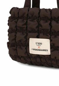 Bolso marrón acolchado hecho de tela suave, con costuras prominentes. Incluye una etiqueta de marca rectangular con detalle de texto.