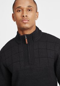 Schwarzer gestrickter Pullover mit einem Reißverschlusskragen, der ein strukturiertes Gittermuster und gerippte Akzente aufweist. Weicher und dicker Stoff.