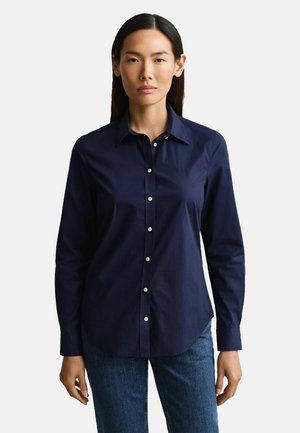 Woman with straight dark hair wears a buttoned navy shirt and blue jeans, standing against a plain light background.
