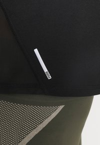 Musta urheilu toppi, jossa on verkkomateriaalia; siinä on pieni valkoinen logo. Alla tummanvihreät legginssit, joissa on geometrinen kuvio ja teksturoitu pinta.