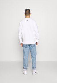 Sudadera de algodón blanca con cuello redondo y puños acanalados, que presenta un pequeño logo de Nike en azul y amarillo en la parte posterior. Jeans de color azul claro y zapatillas blancas.