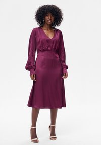 Burgundowa sukienka z satyny z długimi balonowymi rękawami, głębokim dekoltem w kształcie litery V i talią podkreśloną paskiem, sięgająca do połowy łydki. Gładka tekstura i elegancki fason.