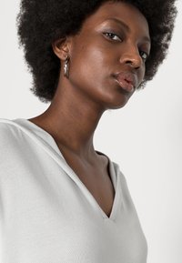Top cinza claro em V com acabamento texturizado e capuz. A modelo usa brincos de argola prateados. Pele lisa e cabelo curto e encaracolado visíveis.