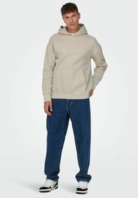 Felpa beige con tasca frontale, caratterizzata da una vestibilità comoda e polsini a costine. Abbinata a jeans di denim blu larghi e sneakers bianco e nero.