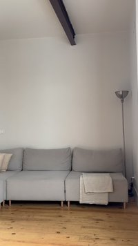 Γκρι καναπές γωνιακός με μαξιλάρια και μπεζ κουβέρτα, ξύλινο πάτωμα, ψηλό ασημί επιδαπέδιο φωτιστικό, λευκοί τοίχοι και εκτεθειμένη δοκός οροφής.