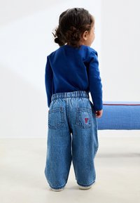 Plava košulja dugih rukava u kombinaciji s širokim, svijetloplavim trapericama s elastičnim pojasevima i crvenom vezenom mašnom na stražnjim džepovima.