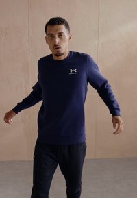 Mörkblå sweatshirt i mjukt tyg, med rund halsringning och ribbade ärmslut, samt en vit Under Armour-logga på vänster bröst.