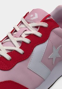 Κοντινή λήψη ροζ και κόκκινου αθλητικού παπουτσιού με λευκά κορδόνια και λογότυπο αστεριού στο πλάι και στη γλώσσα, που δείχνει λεπτομερή ραφή και υφή.