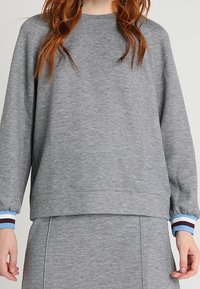 Sudadera gris con cuello redondo, mangas largas y puños acanalados con rayas azules y marrones. Textura suave.