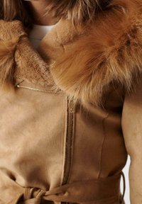 Tan ruskindjakke med en pelskrave, der har en lynlås foran og et bælte i taljen. Teksturen er blød med synlige sømme og glat stof.