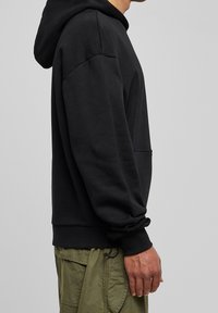 Czarna bluza z kapturem o luźnym kroju, ze ściągaczami na rękawach, kieszenią typu kangur oraz miękką fakturą materiału. Połączona z oliwkowymi spodniami cargo.