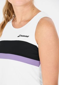 Primo piano di una persona che indossa un top senza maniche bianco con righe orizzontali nere e viola e un logo Babolat sul petto.
