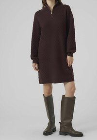 Abito in maglia bordeaux a lunghezza ginocchio, con un motivo a diamante texturizzato, colletto a mezza zip e maniche lunghe ampie; abbinato a stivali alti in pelle marrone.