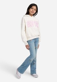 Ragazza che indossa un hoodie beige con la scritta "New York", jeans azzurri con ricami floreali chiari e sneakers bianche, in piedi con una gamba incrociata.