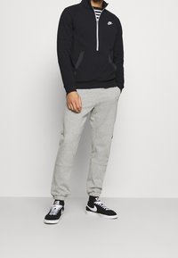 Muž v černém pulovru Nike, šedých joggingových kalhotách a černo-bílých teniskách Nike stojí proti jednobarevnému pozadí.