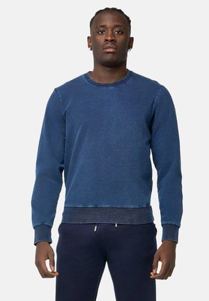 Homme debout face à l'avant portant un sweat-shirt bleu foncé à manches longues et un pantalon bleu marine sur un fond clair uni.