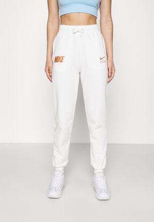 Kobieta w białych spodniach dresowych Nike z pomarańczowymi logo, jasnoniebieskim krótkim topie i białych sneakersach, stojąca na jednolitym tle.