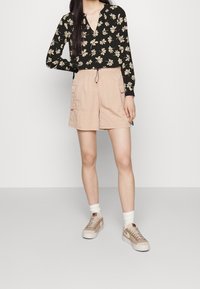Maglietta nera a maniche lunghe con motivo floreale, abbinata a pantaloni cargo rosa chiaro. Il modello indossa sneakers beige con suola bianca e calze.