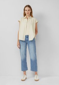 Jasnożółta, krótko rękawowa koszula z guzikami, połączona z niebieskimi spodniami typu wide-leg i beżowymi sneakersami. Miękka tkanina, luźny krój i swobodny design.