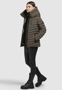 Sieviete valkā brūnu pildītu jaku ar augstu apkakli, melnas legingu bikses un melnas sakstāmas kaujas zābakus, stāvot pretī gaišam fona.