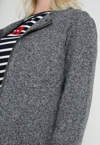 Chaqueta gris texturizada con cierre de cremallera y un sutil patrón tejido, llevada sobre una camisa de rayas con acentos rojos. Vista en primer plano.