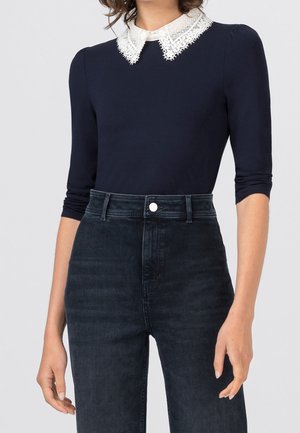 Femme portant un haut bleu marine à manches trois-quarts avec un col en dentelle blanche, assorti à un jean bleu foncé taille haute.