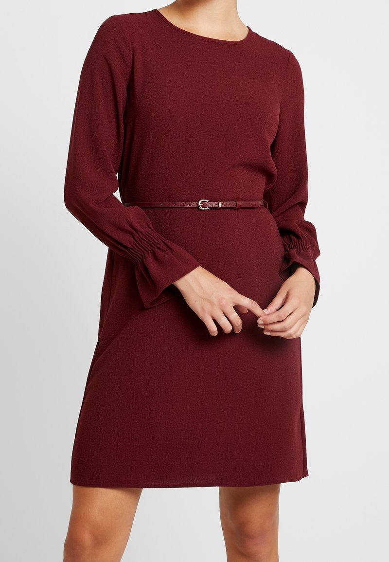 Femme portant une robe bordeaux à manches longues avec une fine ceinture assortie et des poignets froncés, debout devant un fond clair uni.