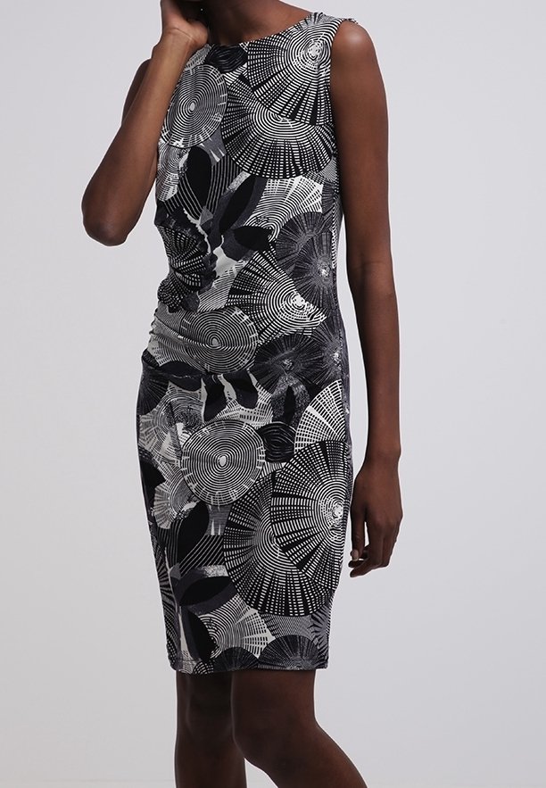 Kvinna som bär en ärmlös, knälång klänning med svarta, vita och grå cirkulära och bladmönster mot en enfärgad grå bakgrund.