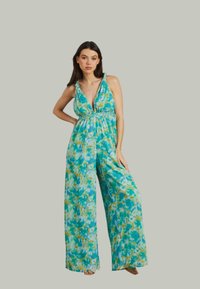 Combinaison tie-dye bleue et verte avec un décolleté halter, taille élastique, jambes larges et des détails minimalistes. Le tissu semble léger et fluide.