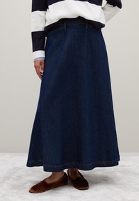 Jupe maxi en denim bleu foncé, avec une coupe droite et des détails de couture subtils, associée à des chaussures à enfiler marron.