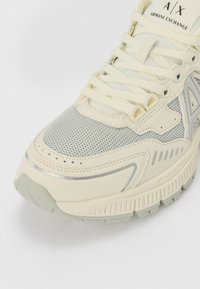 Sneakers gris clair et crème avec des superpositions en maille et en synthétique, bout rond et semelle texturée. Dispose d'un système de laçage et d'une marque en relief.