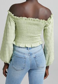 Top verde senza spalline con texture arricciata, scollatura a volant e maniche lunghe, abbinato a jeans aderenti azzurri.