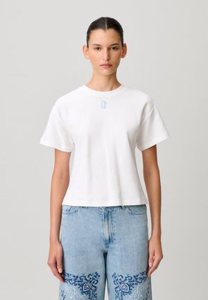 Jeune femme aux cheveux foncés portant une chemise blanche à manches courtes et un jean bleu clair orné de motifs brodés bleu foncé sur les cuisses.