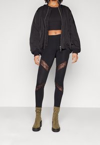 Svart bomberjacka över en svart crop top och leggings med spetsuttal. Accenterad med olivgröna, figurnära ankelstövlar.