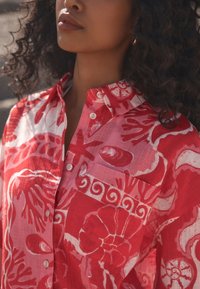 Camicia con bottoni dai motivi floreali rossi e rosa, realizzata in tessuto leggero, con colletto, tasca e motivi bianchi a contrasto.