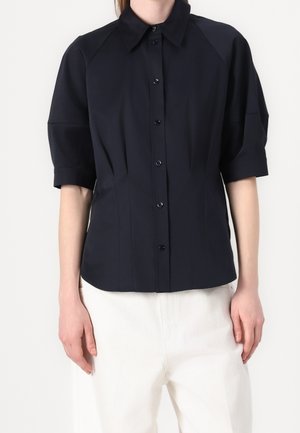 Mujer que lleva una camisa negra de manga corta abotonada y pantalones blancos, de pie con los brazos relajados a los lados contra un fondo liso.