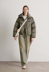 Luftig olivgrønn jakke med høy krage, sammen med lysegrå vide jeans og hvite sneaks. En kremfarget crossbody-veske bæres.