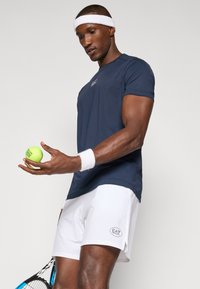 Ναυτικό αθλητικό μπλουζάκι, κοντό μανίκι, με λογότυπο; λευκά αθλητικά σορτς; κρατώντας μια πράσινη μπάλα τένις; λευκός σκούφος και βραχιολάκι συμπληρώνουν την εμφάνιση.