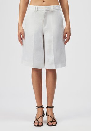 Shorts blancs sur mesure, taille haute et longueur au genou, portés avec des sandales noires à talons hauts et bretelles, sur une personne debout devant un fond uni.